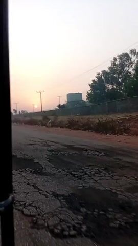 The video shows a road being driven on at dawn or dusk. The road is paved but damaged in some parts. On the right side of the road, there is a low wall and a grassy area with some trees. In the distance, there is a building with a water tank on top. Power lines run parallel to the road. A lone person walks on the side of the road. The sky is a pale pink and orange, indicating that the sun is either rising or setting. The video is shot from inside a moving vehicle.
