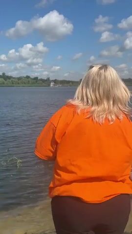A woman with blonde hair wearing an orange t-shirt stands near the edge of a lake. She is looking out at the water. There are two ducks swimming in the lake. The sky is blue with white clouds. Trees are visible in the background. The scene appears to be a park or a natural area.