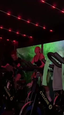 Two women are riding stationary bikes in a dimly lit room. The room has a red light overhead and what appears to be a window covered in green vegetation. The stationary bikes are black with yellow lettering that reads "Cyclone". One woman is wearing a black tank top and the other is wearing a black t-shirt. They are both pedaling at a moderate pace. The room appears to be a gym or fitness studio.