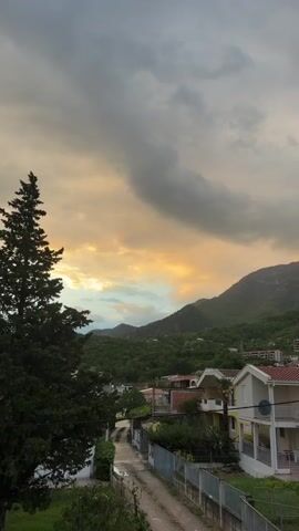 The video presents a scenic pan of a residential area in Zgrade, Montenegro, under a dramatic sky. The scene is dominated by lush green mountains in the background, covered with trees and foliage. In the foreground and mid-ground, several houses with white and light-colored facades, red-tiled roofs, and balconies are visible. Some houses feature satellite dishes and fenced yards with greenery. A dirt path winds between some of the properties. Towards the right side of the frame, a basketball hoop is visible in a yard.

The sky is heavily overcast with large, dark gray clouds, but a prominent band of warm golden and orange light breaks through the clouds on the left side, suggesting either a sunset or sunrise, or the aftermath of a storm. Various trees, including a tall evergreen on the far left and flowering bushes, are scattered throughout the landscape. The overall impression is one of a quiet, mountainous village or town. There are no people or discernible activities taking place, and no visible text.