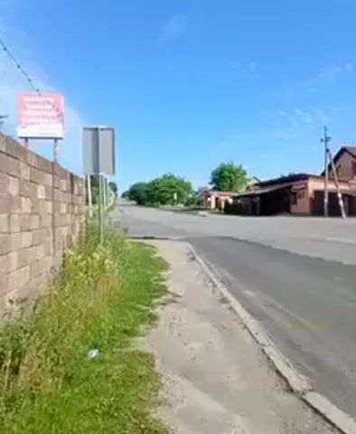 A video pans across a rural road on a sunny day. On the left, a brick wall is visible, followed by overgrown grass and a blue sky. A sign on a pole stands near the wall. Across the road, houses and trees line the street. Further down the road, there are piles of dirt or sand, and agricultural fields in the distance. Cars are parked near one of the houses, and a street lamp is visible. The road appears to be a two-lane asphalt road.