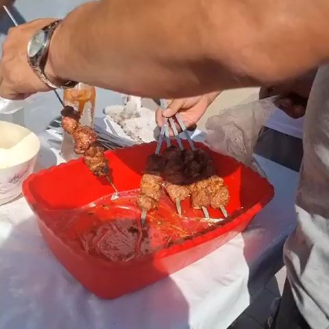 The video shows a close-up view of hands preparing and serving food, likely at an outdoor market or food stall. In the foreground, a person's left arm and wrist, adorned with a watch, is holding a plastic bag containing cooked meat on skewers. The meat appears to be small pieces of seasoned meat grilled to a brown color. The bag is being held open, and the meat is being offered to someone else. In the middle ground, a red plastic container holds several more skewers of meat, some of which are partially submerged in a reddish liquid, possibly marinade or sauce. Behind the red container, a glass bottle with a red cap, containing an amber-colored liquid, is visible. Another person's hands are seen in the background, possibly involved in the food preparation or serving process. The overall setting appears to be outdoors, with a white tablecloth on a table and a blurred background suggesting other market stalls or a street scene. The lighting is bright, indicating it is daytime. The actions suggest a transaction or exchange of food.