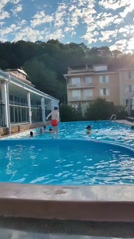 A drone shot shows a group of people swimming in a large outdoor pool. The pool is surrounded by a concrete deck and a tan tiled edge. In the background, there are buildings and a lush green hillside. The sky is blue with some white clouds. The camera pans across the pool, showing people of various ages enjoying themselves. Some are swimming, while others are playing with a float. One person is jumping into the pool from the edge. The drone then moves to the right, offering a wider view of the pool and its surroundings. The sun is setting, casting a warm glow on the scene.