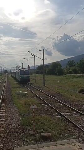 The video shows a Bulgarian State Railways (BDZ) train speeding along a track during daylight. The train is blue and green with a Siemens Smartron locomotive at the front. It is moving on a track with gravel and grass on either side. A small signpost is visible.  In the background, there are trees and mountains under a sky with clouds. The camera remains still throughout the video.