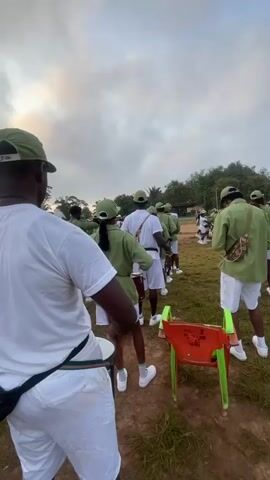 A group of young people in matching green uniforms and white shorts are standing in a line outdoors. They appear to be a marching band, as some are holding drums and drumsticks. The person in the foreground, facing away from the camera, is also wearing a white t-shirt and white shorts, and is holding drumsticks. The sky is overcast.