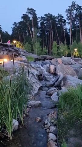 A man and a woman are sitting on a rock formation with lights. A stream flows over rocks, surrounded by grass and trees. There are lights in the trees. The sky is dark.