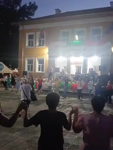 A group of people are dancing in a circle on a paved square in front of a building. The building is yellow with white trim and has a banner across the top that appears to be in a foreign language, possibly Bulgarian, with green and white lettering. There is a Bulgarian flag hanging from the building on the left side. In the background, there is a stage with musicians performing, and several people are watching from the side. There are also tents and tables set up on the left side of the frame, suggesting a festival or outdoor event. The lighting indicates it is dusk or early evening. Some people in the foreground are holding hands and facing away from the camera, also participating in the dancing. The overall atmosphere appears festive and communal.