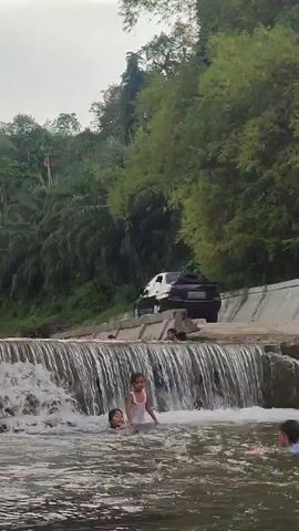 This media file captures a lively scene at a waterfall in Saweuk, Indonesia. The primary subjects are several children playing and swimming in the water. One young girl in a white top stands in the middle of the waterfall, looking towards the camera. Two other children are visible in the water near her, with one partially submerged. In the background, a black car is parked on the road above the waterfall, and several other people can be seen standing on the edge of the waterfall.

The setting is a natural environment with lush green trees and vegetation covering the slopes behind the waterfall. The waterfall itself is a man-made structure, a weir or dam, with water cascading over its edge, creating a dynamic and refreshing atmosphere. The water appears somewhat murky, typical of a natural river.

The activity taking place is recreation and enjoyment of the water, likely a popular spot for families to cool off. The expressions of the children in the water suggest a fun and carefree experience. The time of day appears to be late afternoon or early evening, given the soft, diffused lighting and the overcast sky, which suggests potentially humid or rainy weather common in Indonesia. The overall mood is one of simple, unadulterated fun and outdoor activity. There is no visible text in the media file.