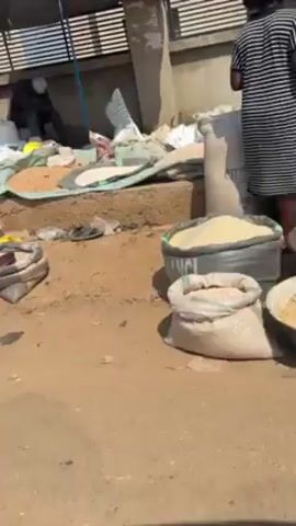 The video depicts a bustling outdoor market scene. Sacks and piles of grains and other goods are spread out on tarps and within a makeshift tent structure. Several people are visible, either standing, squatting, or moving around the goods. The overall environment appears to be dusty or sandy, with simple structures and a fence line in the background. The camera pans slightly, providing a more comprehensive view of the market area.