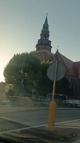 This video captures a view of a historic church in Gryfino, Poland, likely dating back to the 14th century. The camera appears to be mounted on a moving vehicle, possibly a car, given the perspective and the way the foreground is blurred.

The primary subject is the church, with its prominent, multi-tiered steeple and red-tiled roof. The building is constructed with a combination of brick and stone, showcasing Gothic architectural elements like arched windows and decorative stone carvings. Trees and greenery surround parts of the church, suggesting a well-established setting.

In the mid-ground, there are glimpses of street infrastructure, including what appears to be a bus stop shelter and some construction fencing, indicating ongoing work or recent renovations. Several vehicles are visible, including a white car and a bus, suggesting the church is located in a relatively busy area. A few individuals can be seen walking near the church.

The lighting suggests it is either early morning or late afternoon, with the sun casting a strong, warm glow and creating lens flare in the lower left of the frame. The sky is clear and light blue.

A notable detail is a large sign on the side of the church, which appears to be an informational or construction notice. Partially visible text on the sign includes "ST3" and some European Union symbols, possibly indicating funding or a project related to the church's restoration or preservation.

The overall impression is a serene yet active urban environment, with the ancient church standing as a significant landmark. The time of day and the gentle movement of the camera create a peaceful, observational mood.