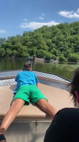 This media file captures a serene moment on a boat ride along the Rijeka Crnojevića river in Montenegro. The scene is set on a sunny day with a bright blue sky dotted with fluffy white clouds, suggesting pleasant weather for outdoor activities.

The primary subjects are two individuals, presumably children, enjoying the boat trip. In the foreground, one child, seen from behind, is lying down on the boat's seating area, wearing a blue t-shirt and green shorts. Their posture suggests relaxation and enjoyment of the surroundings. Another individual, also seen from behind and wearing a dark bucket hat, is seated in the boat, looking out at the river. Their presence indicates a shared experience.

The boat itself is an object of interest, appearing to be a motorboat with a metal railing and a fabric canopy overhead, providing shade from the sun. The boat is moving gently on the water, creating a sense of peaceful travel.

The setting is a picturesque river surrounded by lush green hillsides densely covered with trees. The water is calm and reflects the blue sky and greenery. Patches of lily pads or similar aquatic vegetation dot the surface of the water, adding to the natural beauty of the scene. In the distance, along the riverbank, a small wooden structure and what appears to be a floating platform or a small boat shed are visible, hinting at human presence and activity in this natural environment.

The overall atmosphere conveyed is one of tranquility and leisure. The clear weather and the gentle movement of the boat contribute to a feeling of calm and enjoyment. The focus is on the natural beauty of the location and the simple pleasure of a boat ride. There is no visible text in the media file.