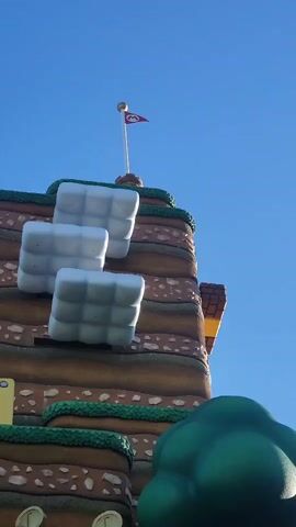A low-angle video showcases a section of a Super Mario-themed attraction. The frame is dominated by a multi-tiered, layered structure resembling a cartoonish earthen landscape, painted in shades of brown, tan, and green. Atop the highest tier, a flag pole with a red flag bearing a white "M" emblem is visible against a clear blue sky. Three oversized, white, cube-like blocks with a grid pattern are attached to the side of the structure. Below these, a yellow block with a diamond pattern sits on a ledge. Further down, a brick-like structure features a yellow and red "POW" block with two gold coins protruding from it, and a dark opening. A spiked gray block with a cartoonish angry face is to the left of the POW block. To the right, a large, stylized green tree stands with a few red mushrooms at its base. Below the tree, a stack of three brown mushroom-like creatures, identified as Goombas from the Mario franchise, are arranged vertically. A large green pipe is positioned in the lower foreground. The overall impression is of a vibrant, playful, and fictional environment.