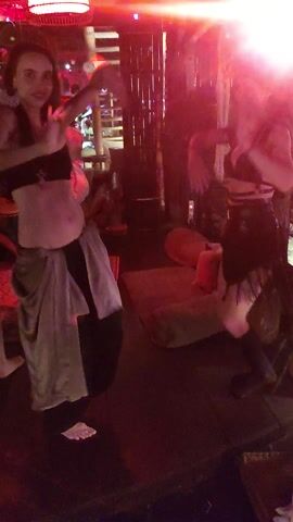 The video captures an energetic night scene inside a club or bar in Lokaserena, Indonesia. The setting is dimly lit, with prominent red and purple party lights creating a dynamic atmosphere and casting a bright red glow on the left side of the frame. The footage is taken from an elevated perspective, looking down at the patrons.

Several people are visible, mostly women, engaged in dancing and socializing. In the foreground, a woman wearing a black top and a light-colored, possibly white or grey, skirt is lying on her back on a raised platform or floor, moving her legs and torso as if dancing. Another woman in a black outfit is standing and dancing nearby. Other individuals are partially visible in the background, some seated and some standing, near what appears to be a bar counter or a similar setup with wooden structures and decorations. The general ambiance is lively and festive. No visible text is present.
