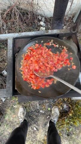 Here is a detailed description of the video:
The video captures a bird's-eye view of someone cooking outdoors. A person is stirring diced tomatoes and onions in a large, flat, cast-iron pan set over a simple metal stove frame. The cook uses a large, slotted metal spoon to mix the ingredients, which are steaming as they cook. The ground around the stove is covered with dry leaves and grass. The person operating the spoon wears black pants and light-colored sneakers. Another person’s feet, clad in flip-flops, are visible on the bottom right of the frame.