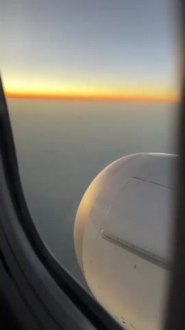 The video shows a view from an airplane window. Outside the window, the sun is setting, casting a warm orange glow on the horizon. Below the horizon, the earth is visible, shrouded in a haze. Part of the airplane's engine is visible in the foreground, its white casing reflecting the sunlight. The window frame is dark and slightly blurred, creating a sense of depth and perspective. The overall impression is one of serene beauty and peaceful travel.