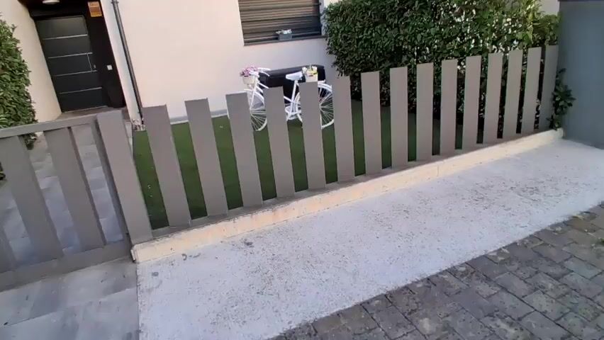 The video pans from left to right across the front yard of a house. A gray fence runs horizontally across the foreground, with a section of gray paver walkway to the left and a paved driveway leading to a dark gray front door on the far left. Behind the fence is a patch of artificial turf. In the center of the artificial turf is a white decorative bicycle with a basket of pink flowers on the handlebars. To the right of the bicycle is a dark storage box. Behind the bicycle and storage box is the exterior wall of the house, which is cream-colored, with a dark-slatted window above. To the right of the house wall is a dense green hedge.