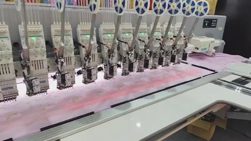 The video shows a wide shot of an industrial embroidery machine in operation. The machine features a long row of multiple embroidery heads, each with a white casing and green indicator lights. The heads are positioned over a long piece of light pink fabric, which is being embroidered with a repeating pattern of small, circular designs in a darker pink color. The fabric is stretched taut and appears to be moving horizontally. The embroidery machine itself is silver and white, with visible metal components and numerous spools of thread at the top of each head. In the background, more rows of similar embroidery heads are visible, stretching into the distance. To the far left, a person in a dark suit and a white shirt is partially visible, standing near a display with shelves. The environment appears to be a factory floor.