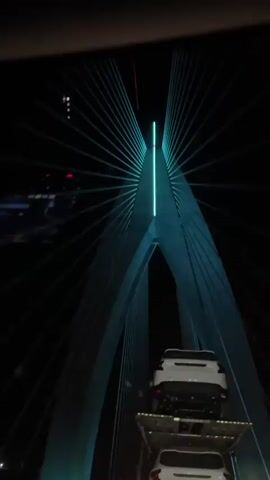 From the perspective of a vehicle, the camera films two white cars stacked on a trailer driving across a bridge at night. The bridge has a large concrete structure lit with bright blue lights. The bridge's cables appear white against the night sky. The road is brightly lit.