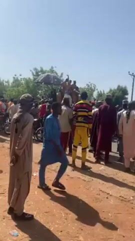 The video shows a crowd of people gathered on a dirt road under a clear, sunny sky. Many of the people are wearing traditional clothing. In the background, some individuals are on an elevated platform under a dark umbrella. There are motorcycles parked along the road. The people appear to be observing the scene, and some are moving around.