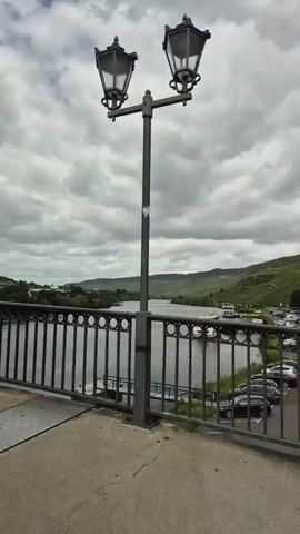 A first-person view from inside a moving car, looking out the front window and then panning to the right to show the scenery through the passenger window. The car is driving on a bridge over a wide river. On the other side of the river, there is a town with buildings and a parking lot filled with cars. Rolling hills covered in green vegetation, possibly vineyards, are visible in the background under a cloudy sky. The car then drives over the bridge, showing an oncoming black sedan and other vehicles.