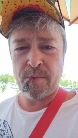 A man is taking a selfie outdoors. He is wearing a white t-shirt and a novelty hat shaped like a donut. He has a cigar in his mouth and is looking at the camera. Behind him, there is an airport terminal building with people and luggage. He is holding a red strap, possibly from a bag or a camera.