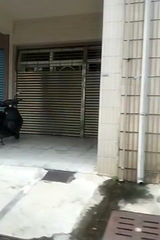 The video pans across a street scene with several closed storefronts. A dark-colored scooter is parked in front of a blue rolling shutter. Another scooter, white with a license plate, is parked further down. A potted plant is visible to the left. The ground is tiled and appears wet in places. There are some pipes and a yellow hose on the right side of the frame. A small ramp is placed in front of one of the shutters.