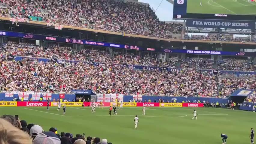 The video shows a soccer match being played in a large stadium filled with spectators. The field is green, and the players are wearing white and dark blue uniforms. The crowd is cheering and appears to be engaged in the game. There are also advertisements and banners visible around the stadium, including one that says "FIFA WORLD CUP 2026". The lighting suggests it is daytime, possibly late afternoon or early evening. The camera is positioned in the stands, looking down at the field from a slight angle. The action on the field is focused on a group of players near the center of the pitch.