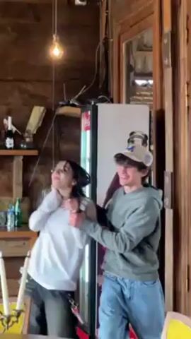 A young woman and a young man are in a rustic room and dancing. The woman wears a white top and black pants and has dark hair tied with a yellow flower. The man wears a beige baseball cap, a grey hoodie, and blue jeans. He guides the woman’s arms as they dance. Behind them is a wood-paneled wall with a shelf holding liquor bottles and glassware. There is also a refrigerator with a Coca-Cola advertisement. The room has wooden furniture and a light fixture hanging from the ceiling.