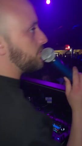 The video shows a DJ in a crowded nightclub setting. The DJ, a man with a beard, is holding a blue microphone and appears to be speaking into it. He is positioned behind a DJ mixer with various controls and buttons. The foreground is filled with the DJ equipment and cables. The background shows a large crowd of people, mostly dressed in white shirts. The nightclub is illuminated with colorful lights, including blue and purple hues. Balloons, possibly red, are seen hanging above the crowd. The overall atmosphere is energetic and festive.