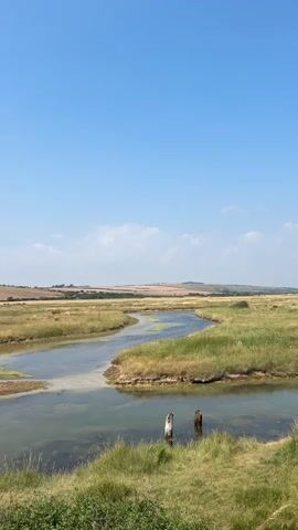 The video shows a rural landscape with a winding river or stream flowing through a grassy marshland under a clear blue sky with a few scattered clouds. In the foreground, there are posts in the water, and the path leads to a person walking away from the camera along a paved path beside a body of water. The landscape appears to be dry and sunny, suggesting a warm day.