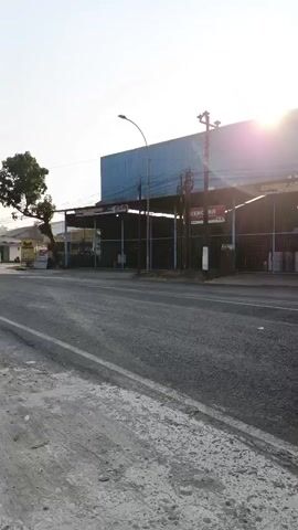 The video shows a roadside view with a street in the foreground and a building with a large blue awning in the background. The sun is low in the sky, casting a lens flare across the scene. A black SUV drives away from the camera on the right side of the road. A red motorcycle with a rider wearing a helmet and a backpack approaches the camera from the left and passes by. The rider is wearing a light-colored jacket. The road appears to be asphalt with some debris on the shoulder. There are some trees on the left side of the road. The building in the background seems to be a commercial establishment, possibly a warehouse or a retail store, with open bays. There are some traffic cones visible on the right side of the road.