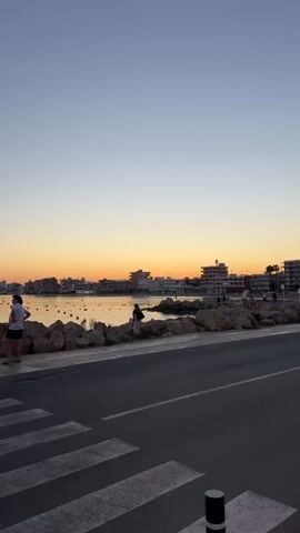 The video shows a tranquil coastal scene at sunset, with a clear, gradient sky transitioning from light blue to orange at the horizon. The camera captures a low angle across a road with a zebra crossing, which leads to a rocky shoreline. Along the shore, various people are observed, some standing alone and others in groups, enjoying the view. They are mostly silhouetted against the setting sun. The water is calm, reflecting the colors of the sky, and in the distance, a cityscape is visible along the horizon. A few boats are visible on the water. The camera slowly pans along the shore and people, ending on a signpost. The scene evokes a sense of peace and leisure.