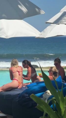 The video depicts four women lounging by a pool overlooking the ocean. They are all wearing swimwear. Two are seated on the edge of the pool, while the others relax on cushioned seating. One woman is applying sunscreen to the back of another. Drinks are visible, and beach umbrellas provide shade. The ocean behind the pool has noticeable waves. The sky is clear and sunny.