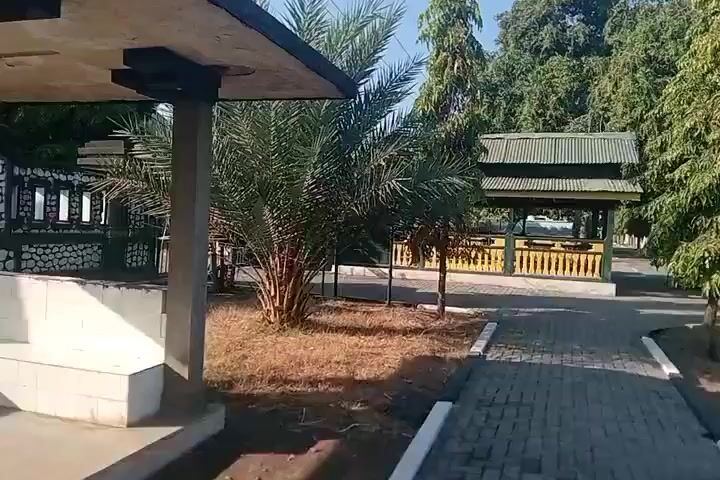 The video shows a paved walkway leading through an outdoor area with several trees and structures. On the left, there is a covered structure with a flat roof and columns. In the center, there is a yellow and green gazebo-like structure with a sloped roof, surrounded by several trees. To the right, there is a fence and a dark-colored monument with a red and green ribbon. The ground is covered with dry leaves and dirt. The sky is clear and blue. The time is daytime.