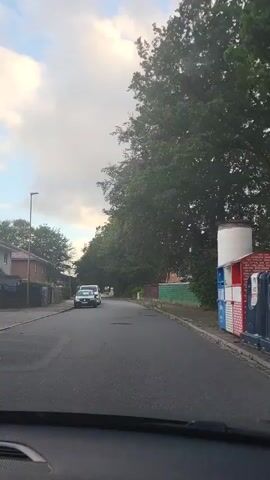 This is a first-person perspective video taken from inside a vehicle as it drives down a residential street. The street is paved and appears to be a two-lane road. On the left side of the road, there are houses with lawns and fences. Several cars are parked along the side of the road, including a white van. On the right side of the road, there are trees and some fencing. The sky is cloudy with patches of blue. The overall environment appears to be a suburban neighborhood.