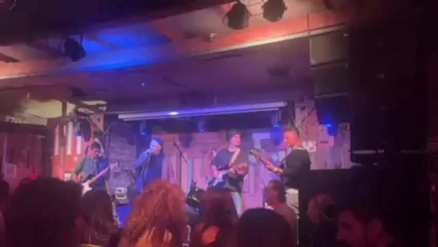 The video shows a band performing on a stage in a dimly lit venue.  The band has five members: a guitarist on the left, a singer in the middle left, a guitarist in the middle, a bassist on the right, and a drummer behind them, though the drummer is mostly obscured.  The stage has a rustic wooden backdrop with a white projection screen above the musicians. There are blue and purple stage lights. The audience is visible in the foreground, mostly from the shoulders up, and they appear to be watching the band. A large stack of speakers is on the right side of the frame. The ceiling is exposed wood beams with various lights and fixtures.