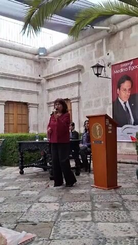 A woman with brown hair, wearing a maroon blouse and black pants, stands at a wooden podium and speaks into a microphone. She gestures with her right hand as she speaks. Behind her, a banner displays the name "Luzgardo Medina Egoavil" with a portrait of a man in a suit and tie. To the left of the podium is a decorative black table with a water bottle on it. Two people are seated behind the table. The scene is set in an outdoor courtyard with stone paving and surrounding buildings. Palm fronds are visible overhead, suggesting a warm climate.