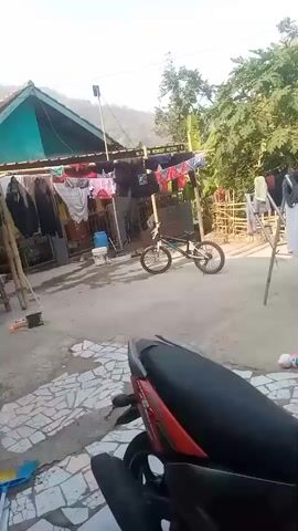 The video shows an outdoor scene in what appears to be a rural or semi-rural area. In the foreground, the back of a red and black motorcycle is visible. Beyond the motorcycle, there is a paved or concrete ground area with a patterned tile section. In the middle ground, a BMX bicycle is parked on the ground. Clothes are hanging on a clothesline stretched between bamboo poles. Behind the clothesline, there are structures that look like simple houses or sheds with green roofs. Trees and vegetation are visible in the background, and a hillside can be seen further in the distance. The sky is clear and blue, suggesting a sunny day. The overall impression is one of a domestic, everyday setting, possibly a yard or courtyard.