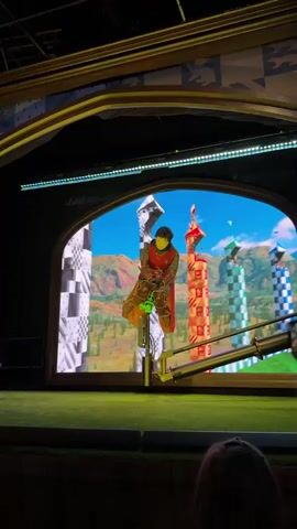 The video captures a simulated Quidditch match. A performer, dressed in a Hogwarts uniform, is positioned on a mechanical broomstick. The broomstick is attached to a rig that allows for movement, simulating flight. The backdrop is a large screen displaying a Quidditch pitch with colorful goal posts and a mountainous landscape under a bright blue sky. The performer's movements are synchronized with the visuals on the screen. The foreground shows a dimly lit audience area with the backs of several heads visible, suggesting the video was taken from within a theater or attraction. Green turf is present in the immediate foreground.