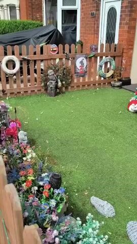 The video shows a small, artificial grass yard filled with garden ornaments. On the left, a wooden fence separates the yard from a building. The fence has several decorative items attached to it, including a circular life preserver, a poster of a witch, and a carved wooden owl. The yard is decorated with many gnome figurines, stuffed animals, and other whimsical garden decorations. There are also two large shell-shaped rocks on the grass. In the center of the yard, there are a series of eagle sculptures perched on a trunk, surrounded by flowers and more gnome figures. To the right, there is a tall giraffe statue and a large orange drink-shaped planter. A rooster sculpture is visible on a wooden fence in the background. The scene is brightly lit, suggesting it is daytime.