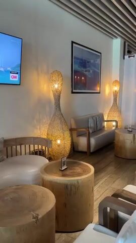 The video pans across a well-lit, modern lounge area. In the foreground, there are several light-colored, plush chairs with wooden frames, arranged around low, round wooden tables. Behind these, two tall, woven lamps with a warm glow stand flanking a cream-colored sofa with a wooden frame. The walls are painted a neutral tone, and a framed piece of artwork hangs on one wall. Further into the lounge, white sheer curtains create partitions, and more wooden tables and chairs are visible. The ceiling features a slatted wooden design with recessed lighting. The camera then moves towards a buffet area, which has a long wooden counter with various serving dishes, a beverage dispenser, and stacked plates and bowls on shelves below. The flooring throughout the space is made of light-colored wood planks. No people are visible in the video.