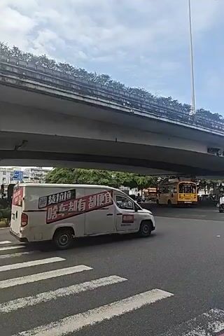 The video shows a street scene under an overpass. A white van with text on the side is driving away from the camera. A white car is stopped at a crosswalk. More cars and scooters are visible in the background, along with trees and buildings. The sky is cloudy.