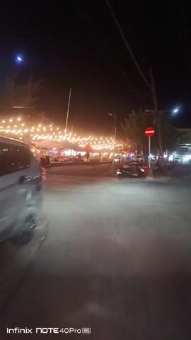 A nighttime shot shows a bustling street market with numerous stalls and vendors. String lights illuminate the area, casting a warm glow on the scene. People can be seen walking around, browsing the stalls, and enjoying the atmosphere. The camera pans from left to right, capturing the vibrant energy of the market. The overall impression is one of a lively and inviting place to be.