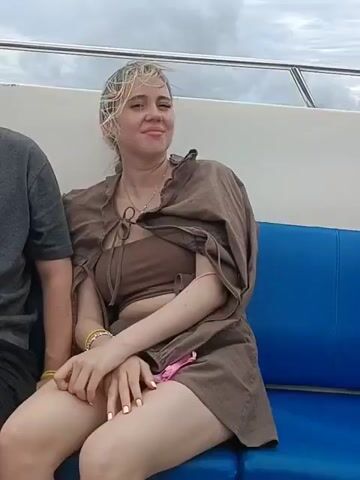A young woman with blonde hair is sitting on a boat, looking to the side with a slight smile. She is wearing a brown bikini top and a brown, loose-fitting shirt draped over her shoulders. Her hands are clasped in her lap, and she has bracelets on her wrists. The boat has blue seating and a metal railing. In the background, there is a body of water and hills under a cloudy sky. The camera pans slightly to show more of the sea and the distant hills. Another person is visible to the right of the woman, wearing glasses and a blue and white striped shirt. The video continues to show the water and the landscape.