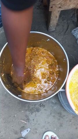 A person is mixing a golden-colored stew-like substance in a large silver metal pot. They are using their bare hand to stir the contents, which appear to contain visible ingredients. The pot is sitting on a concrete surface. Next to it is a round, yellow, mesh colander. The person is wearing a black shirt and jeans, and part of their foot is visible, showing a white sandal. There's also a wooden bench nearby.