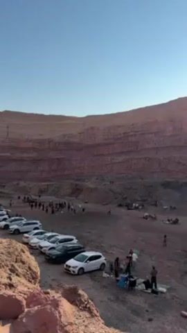 The video shows a group of people gathered in what appears to be an old quarry or a similar excavation site with layered rock walls. A line of parked cars is visible on the edge of the quarry, and people are milling around, some seemingly picnicking or setting up areas to relax. There's music playing, creating a festive or relaxed atmosphere. The quarry walls are a reddish-brown color, and the sky is clear and blue, suggesting it is daytime.