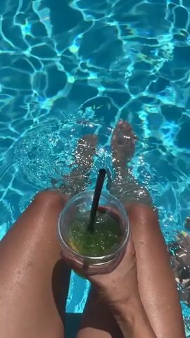 The video opens with a close-up view of a person's legs submerged in a bright blue swimming pool. The camera pans slightly to reveal a clear plastic cup filled with a green beverage and a black straw, held in the person's right hand. Another person's feet, with yellow nail polish, are visible in the lower right corner of the frame, also in the water. The sunlight creates shimmering patterns on the surface of the water. The overall scene conveys a relaxed and summery atmosphere.