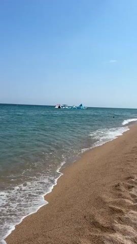 The video shows a sunny day at a beach in Blanes, Spain. The scene is set by the turquoise ocean and a sandy shore. In the distance, a water park with inflatable slides and structures is visible. There are a few people enjoying the water park and some swimming in the sea. The sky is clear and blue, suggesting pleasant weather. The overall atmosphere is one of a relaxed summer day at the beach, with opportunities for recreation and enjoyment in the water. The time of day appears to be late morning or early afternoon, judging by the bright sunlight and clear visibility. No text is visible in the video.