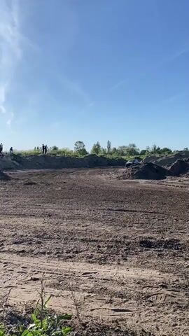 The video shows a black rally car driving on a dirt track on a clear day. The car is labeled with the number 107. It kicks up a lot of dust as it maneuvers around the track. Several people stand on a dirt mound watching the car, and there's green vegetation around the edges of the track. The sky is clear blue with a few wispy clouds.