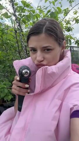 A woman stands outdoors holding a power drill in her hand. She is wearing a pink puffer jacket over a purple shirt. She has light skin, brown hair pulled back, and green eyes. She makes a kissing face, then stops the drill, and then smiles. The background features lush green foliage, a cloudy sky, and a glimpse of a greenhouse. There's also a swing behind her and a pink backpack on the seat of the swing.
